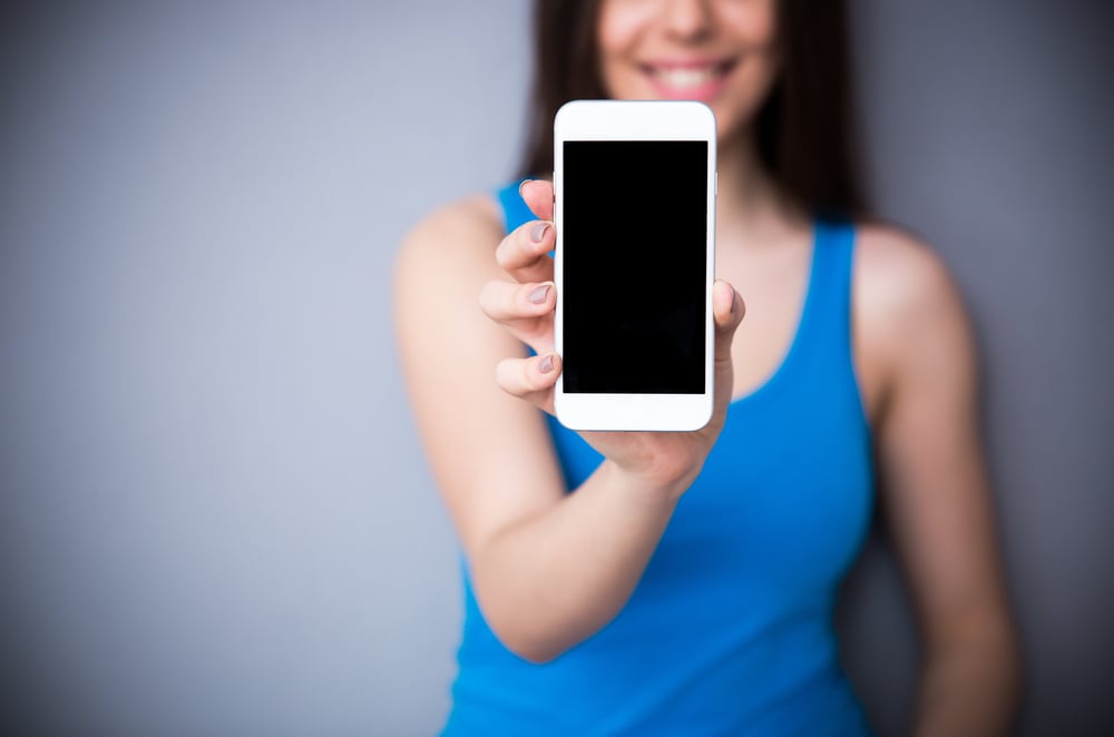 Happy woman showing blank smartphone screen over gray background. Focus on smartphone. Happy woman showing blank smartphone screen over gray background. Focus on smartphone.